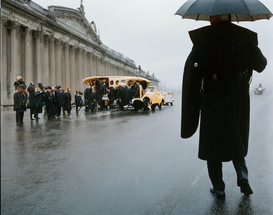 Person with umbrella on rainy day watching people board vintage bus near classical building