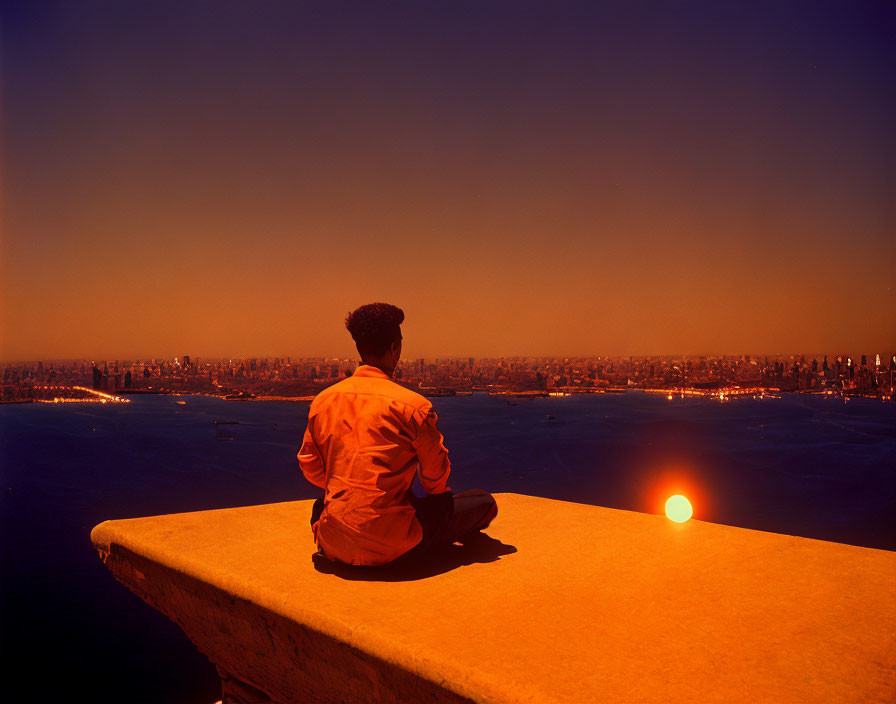 Person sitting on high ledge at vibrant sunset overlooking cityscape and water