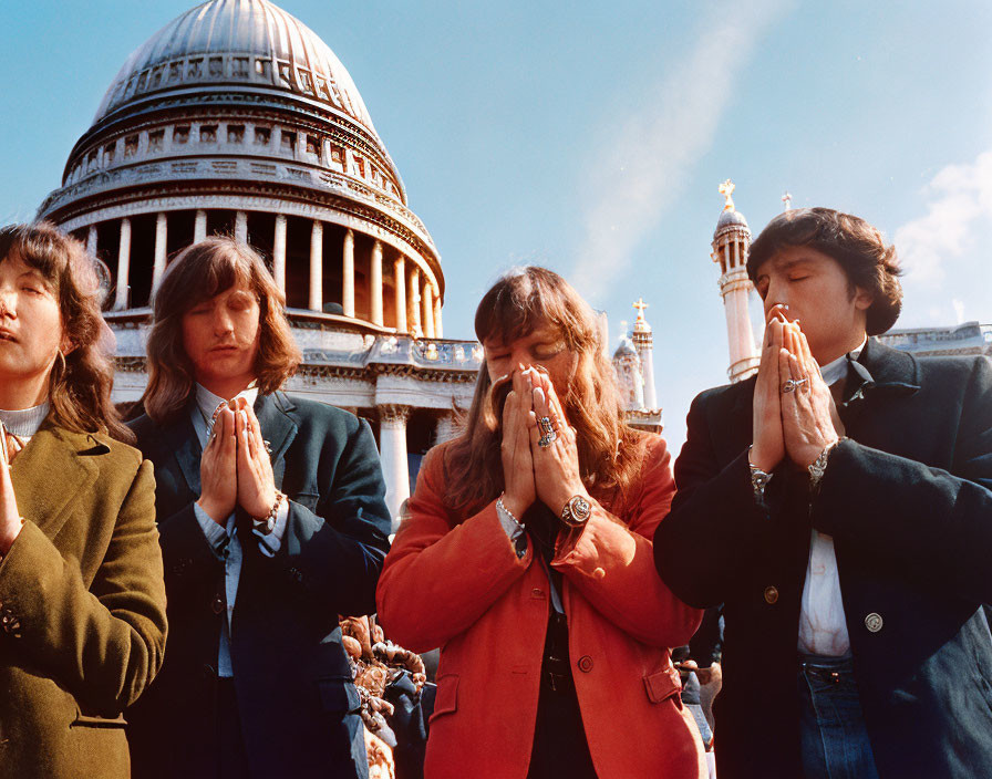 Four people praying with clasped hands, a domed building, and blue sky.