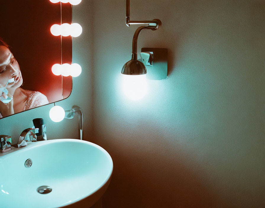 Person applying facial product in mirror with vanity lights by wall-mounted sink under soft lighting