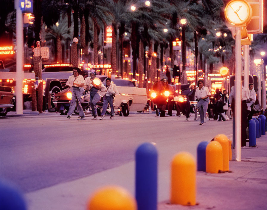 Pedestrians crossing palm-lined street at dusk with cars and clock post under city lights.