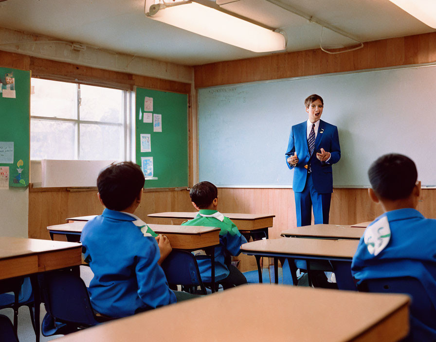 Male teacher in blue suit instructing students in blue uniforms in classroom with desks, whiteboard, and