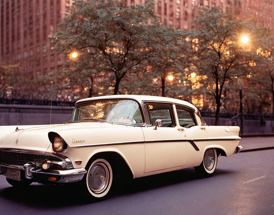 Classic sedan parked on city street at dusk with autumn trees and warm street lights.