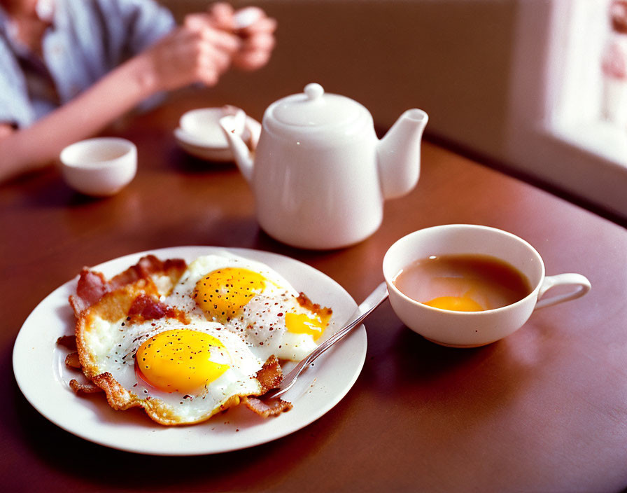 Classic breakfast scene with sunny-side-up eggs, bacon, tea cup, and teapot.