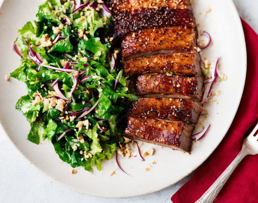 Glazed pork belly with green salad, nuts, and red onions on white plate