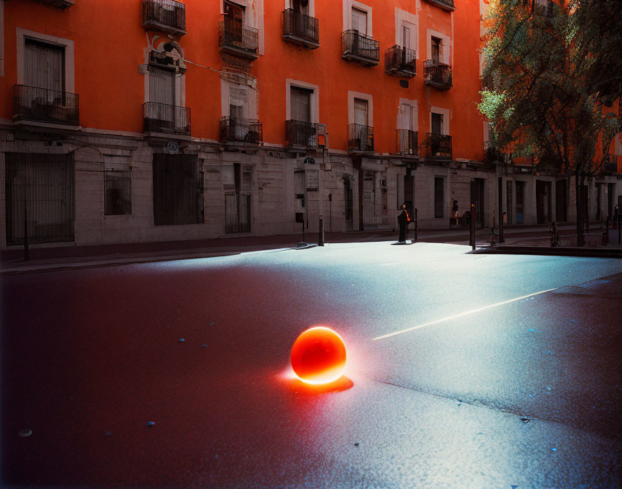 Red glowing sphere on urban street at dusk with reflection, orange buildings and trees in background