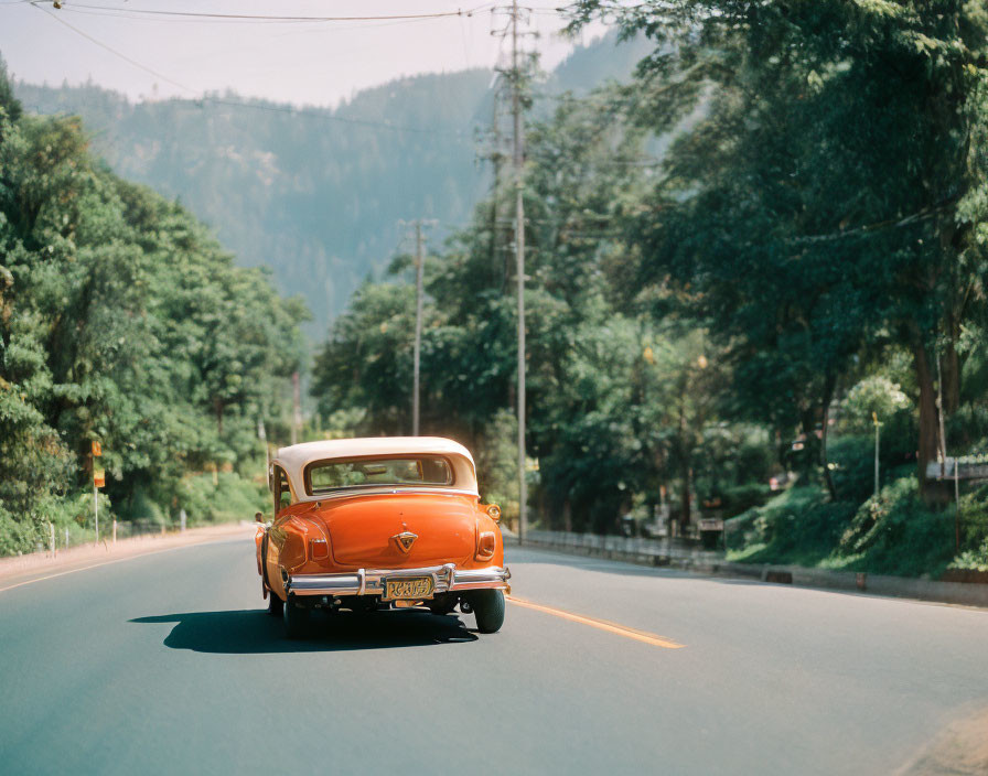 Vintage Orange Car Driving on Empty Road Amid Greenery & Mountains