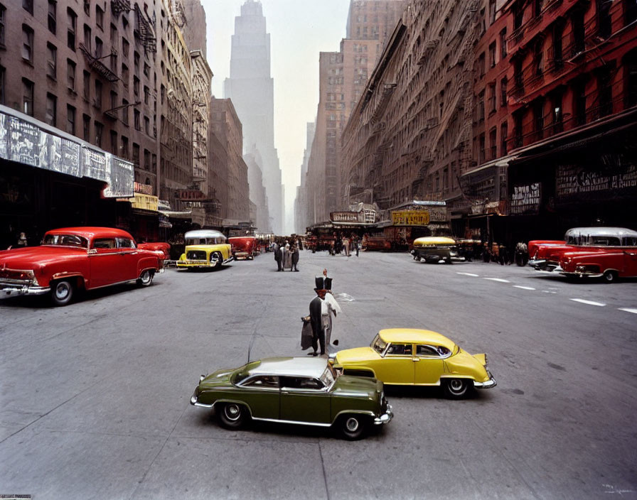 Vintage city street with classic cars and pedestrians amidst towering buildings.