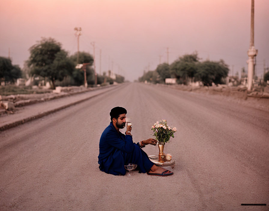 Man in traditional attire sits on deserted road at dusk with pot of flowers and cup.