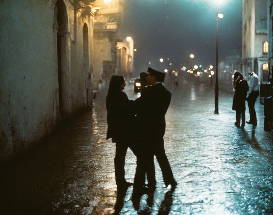 Couple embracing on rain-slicked street at night with street lights and pedestrians.