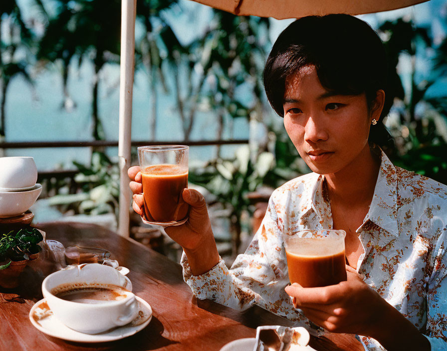 Woman holding tea glass at outdoor table with teapot and cups under white umbrella.