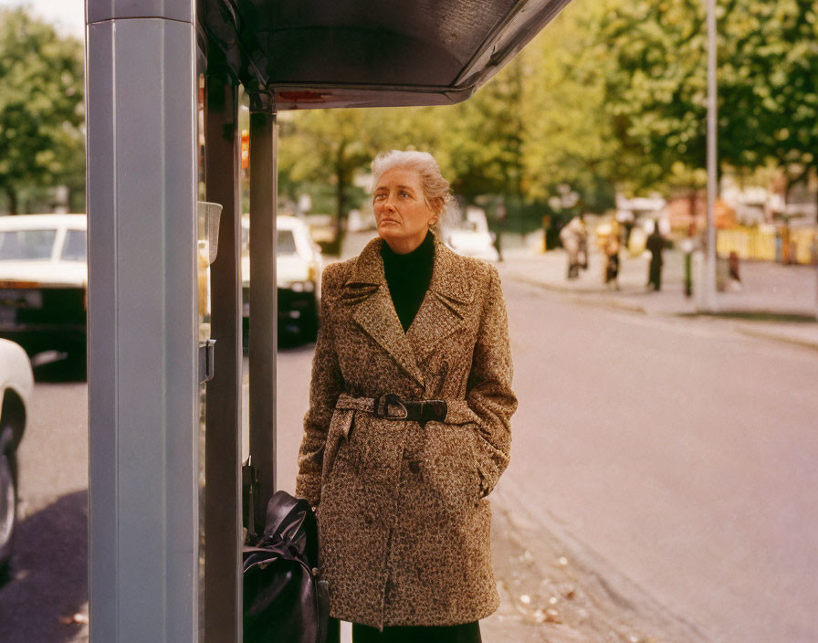 Elderly woman in tweed coat at bus stop with cars and trees.