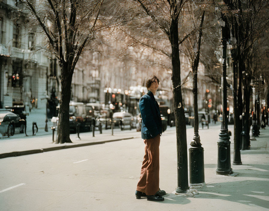 Person standing on sunlit city sidewalk with trees, facing away from camera.