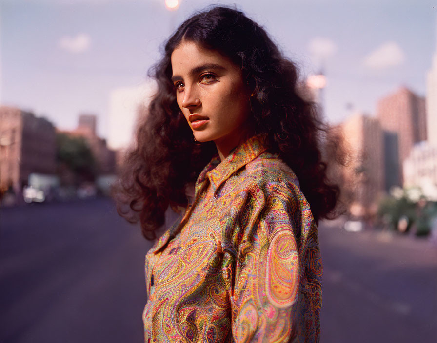 Curly-haired woman in patterned shirt on city street with buildings and blue sky