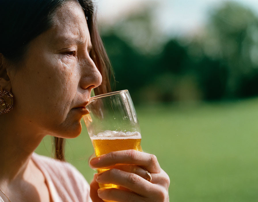 Woman sipping beer outdoors with thoughtful expression and greenery background