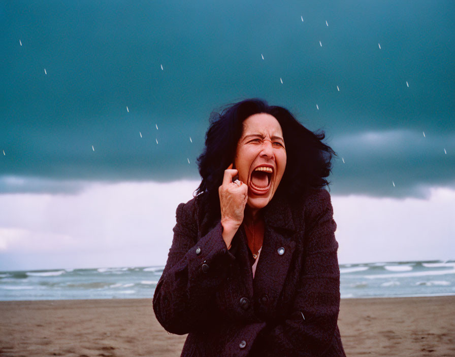 Woman in dark coat screams at beach under stormy sky with snowflakes.