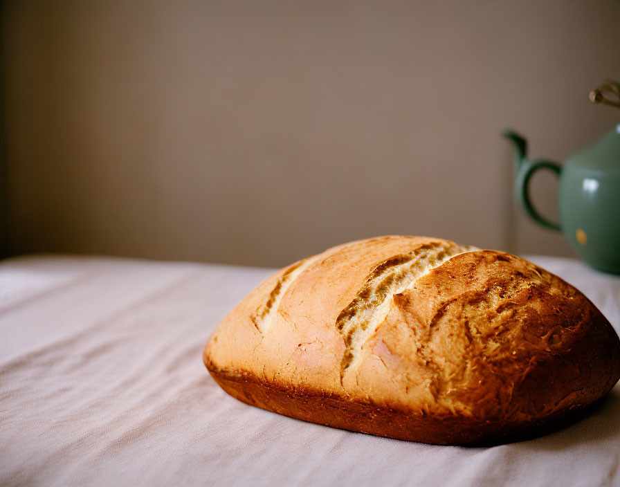 Freshly Baked Bread Loaf on Cloth-Covered Table with Green Teapot