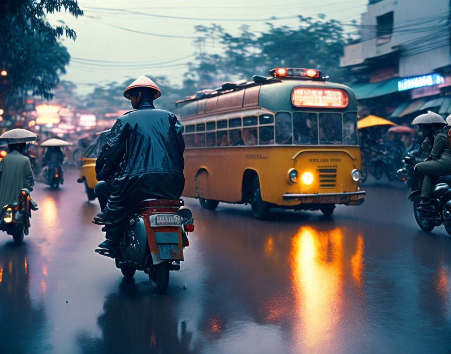 Motorcyclist in raincoat rides near yellow bus on wet street at twilight