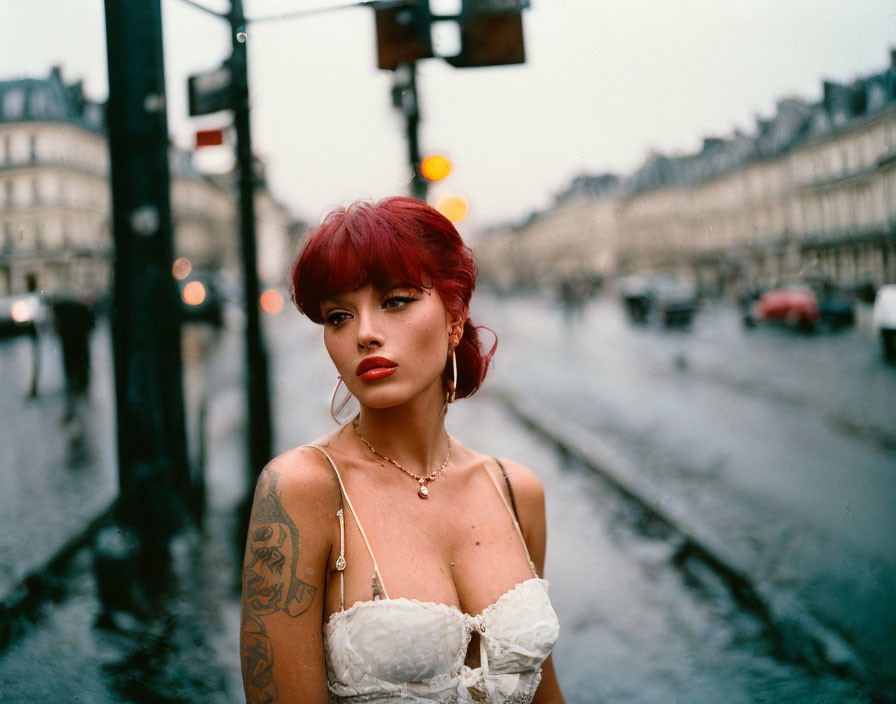 Red-haired woman with tattoos on wet street, white top, blurred city lights.