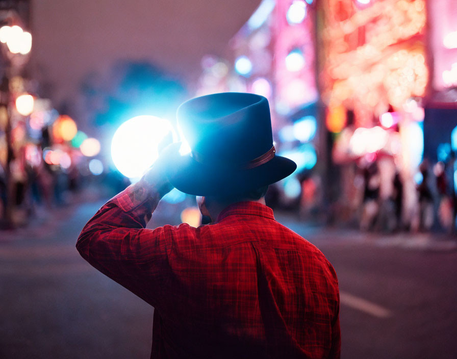 Person in plaid shirt and hat at night, facing away, city lights in background