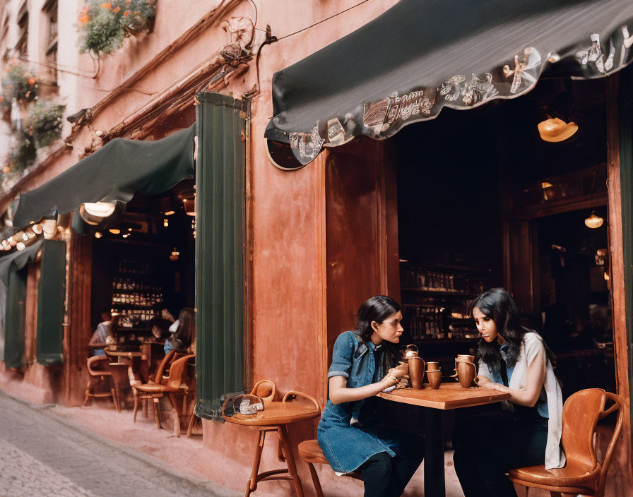 Women chatting at cozy outdoor café with wooden chairs & warm lighting.