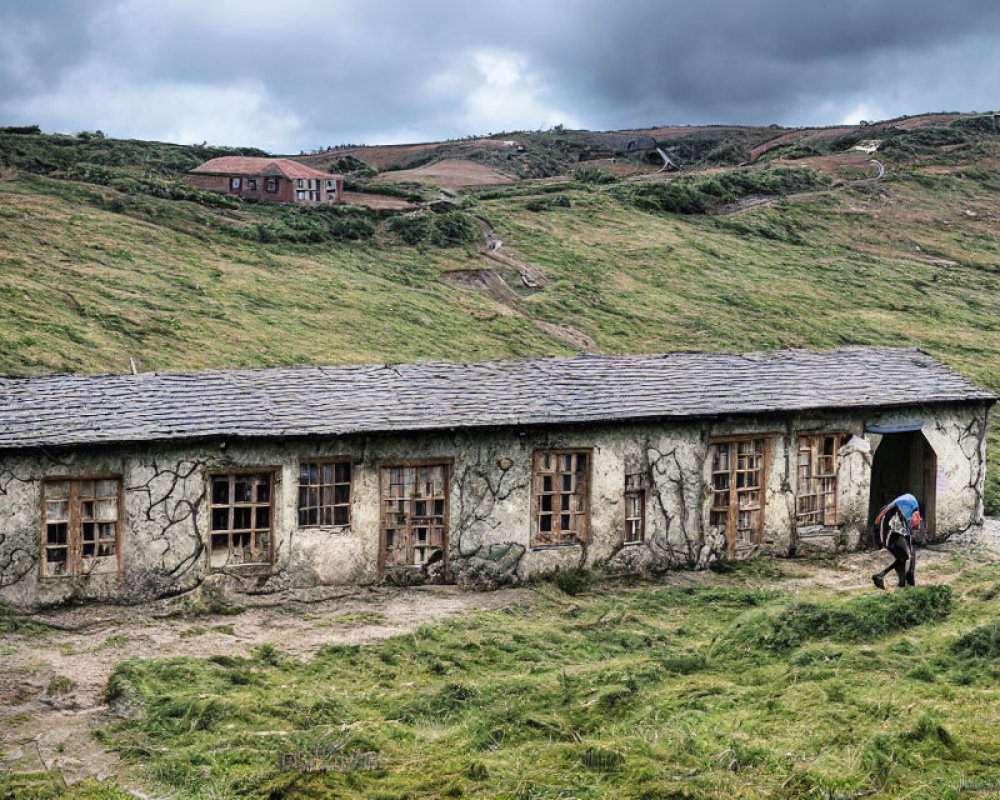 Hiker passing by stone building, grassy hills, and distant house under cloudy sky