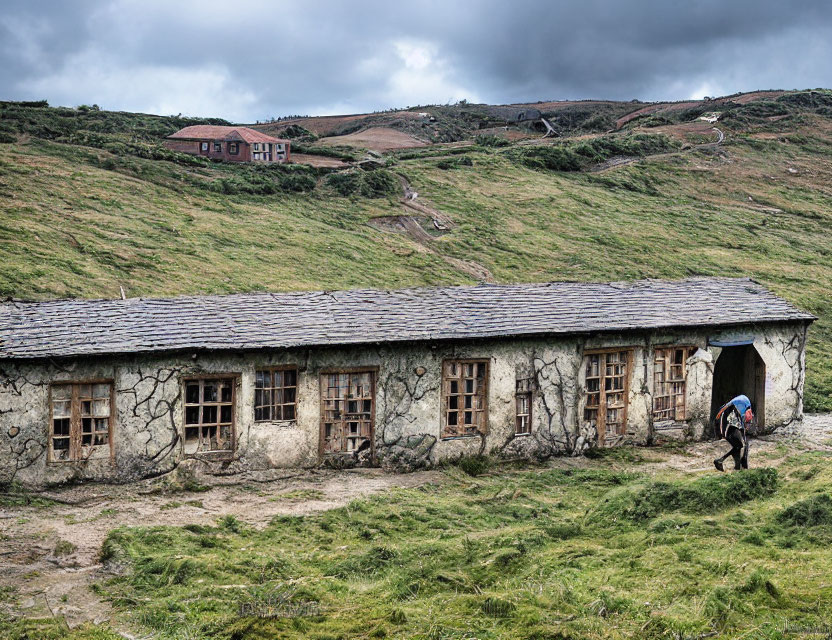 Hiker passing by stone building, grassy hills, and distant house under cloudy sky