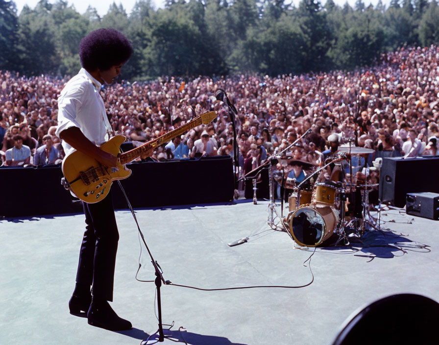 Afro musician plays yellow electric guitar on stage before dense outdoor crowd