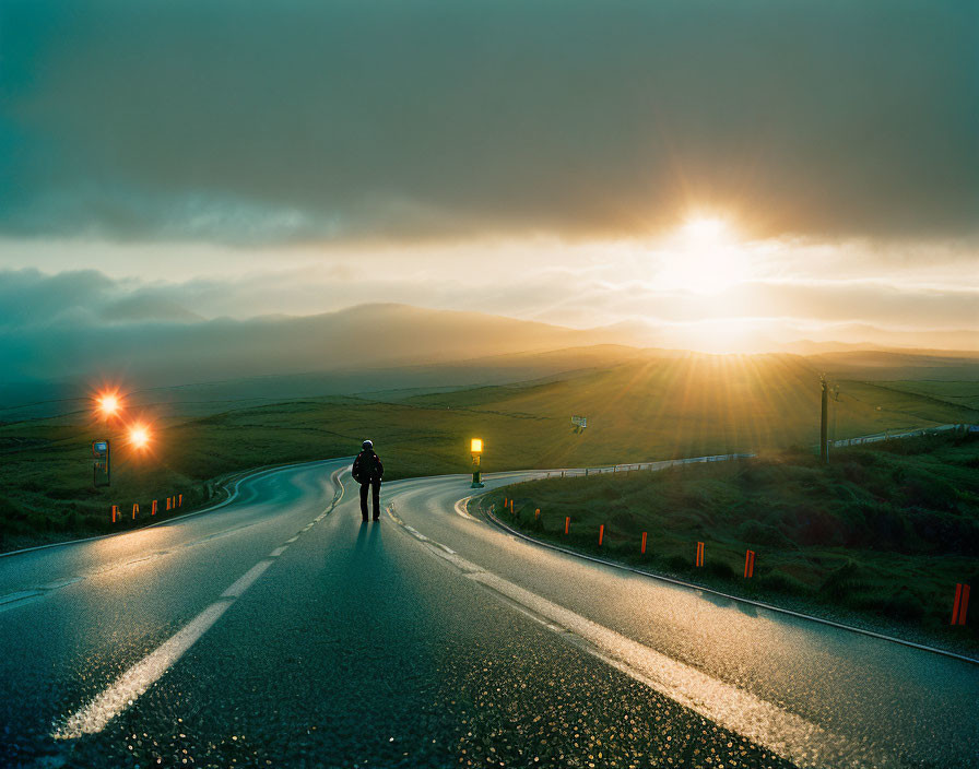 Person standing on curvy road at sunrise with misty hills and sun rays.