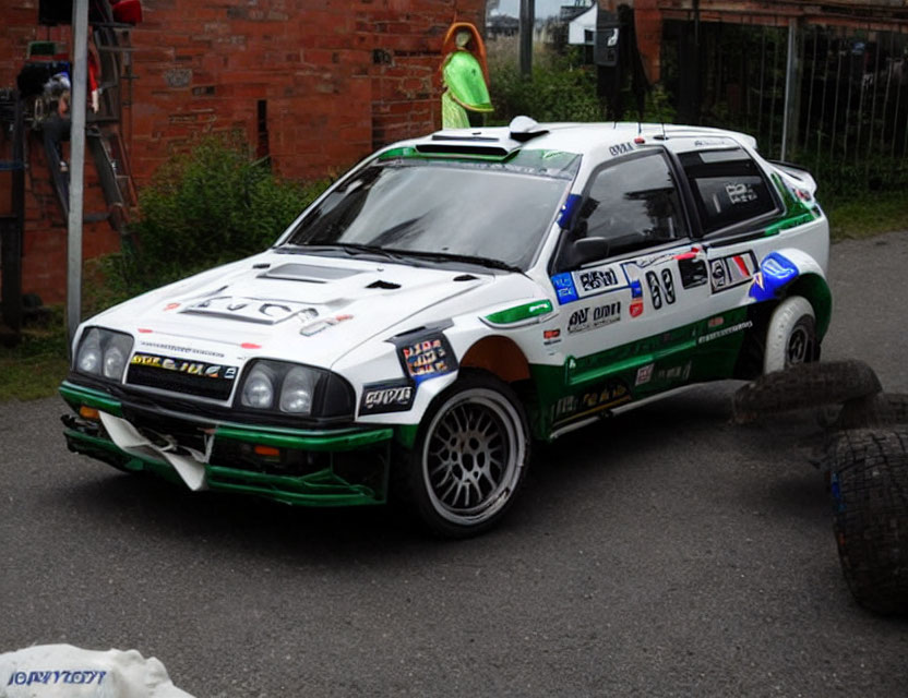 Vintage rally car with green and blue sponsor decals parked on road with person in high-visibility vest.