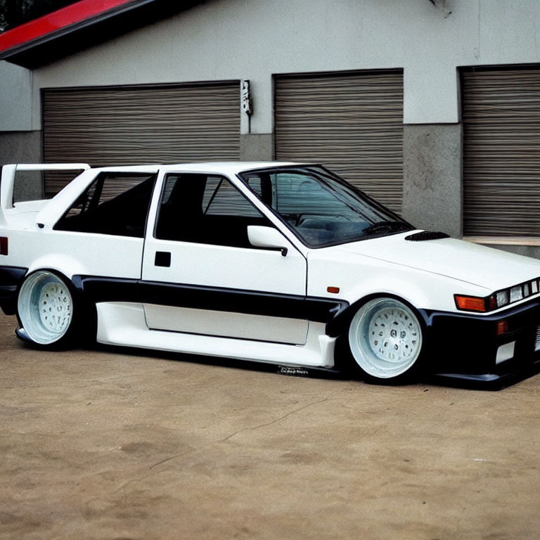 White Vintage Sports Car with Aftermarket Body Kit and Wide Wheels Parked in Front of Industrial Garage Door