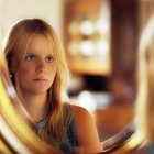 Red-haired woman reflected in round mirror with lamp and another woman's reflection in warm tones