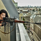 Female sniper in camo pants aiming rifle from high vantage point.