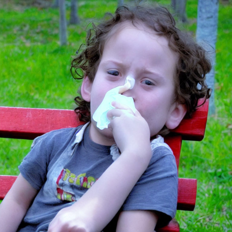 Curly-Haired Child Sitting on Red Bench with Tissue and Frown