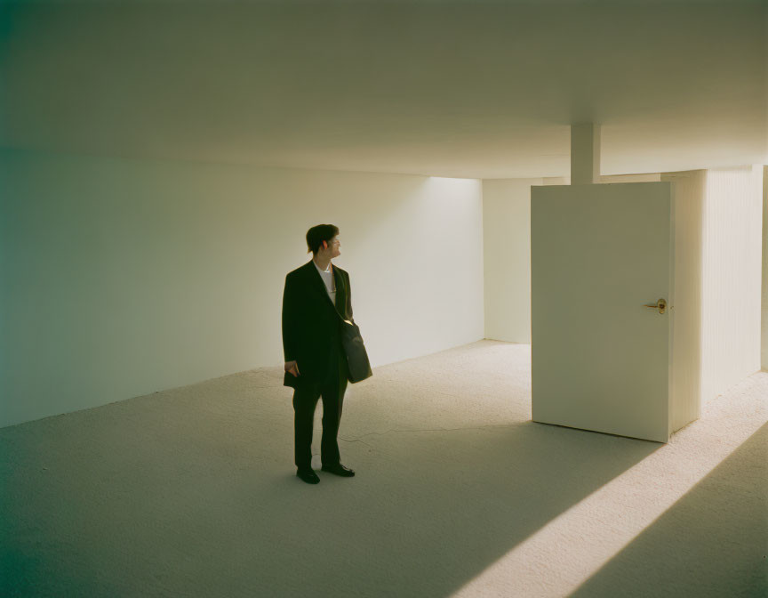 Person in dark suit in sparse room with white walls and shadows.