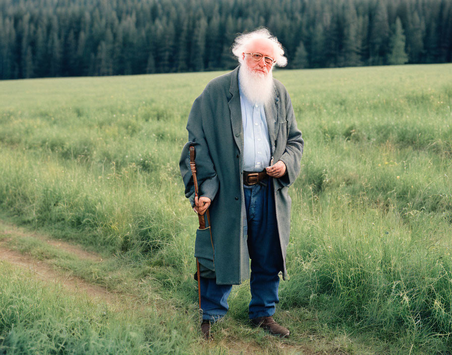 White-bearded elderly man in glasses, hat, and cane in green field with trees.