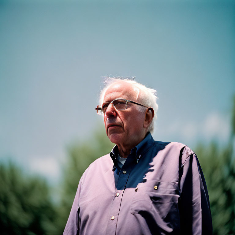 Elderly man with glasses and white hair in blue shirt against cloudy sky.