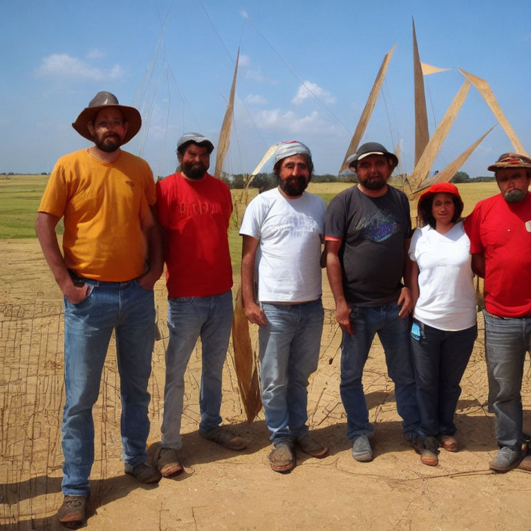 Six people posing outdoors with a large wooden structure under a clear sky