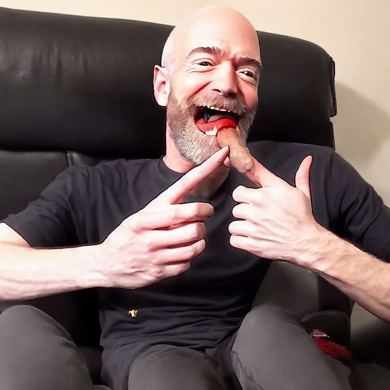 Bald man in black shirt pointing at tongue with spicy snack, laughing