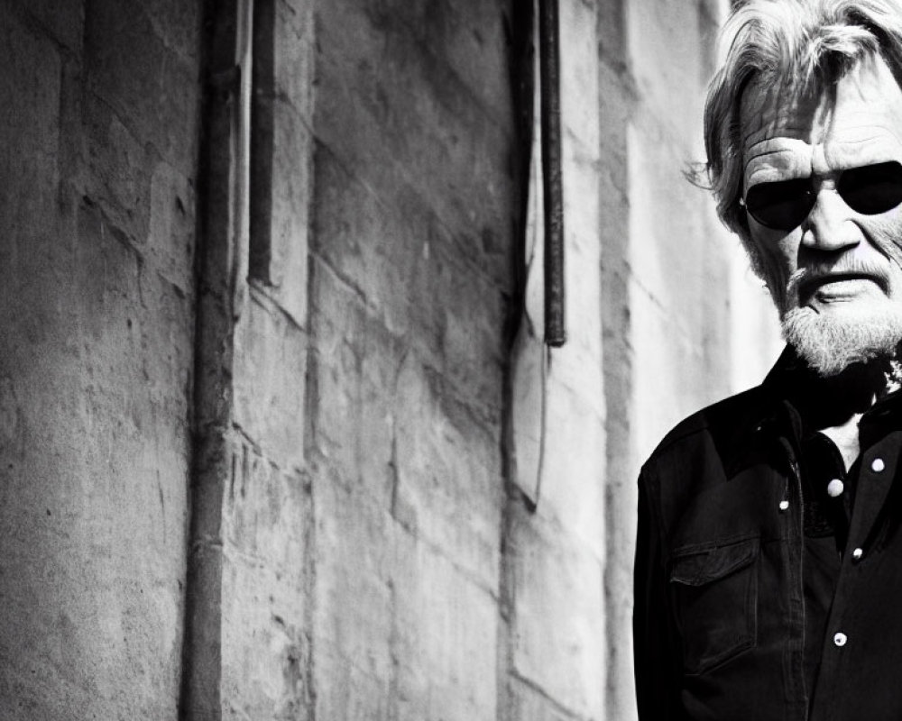 Elderly man in sunglasses with tousled hair standing in front of stone wall