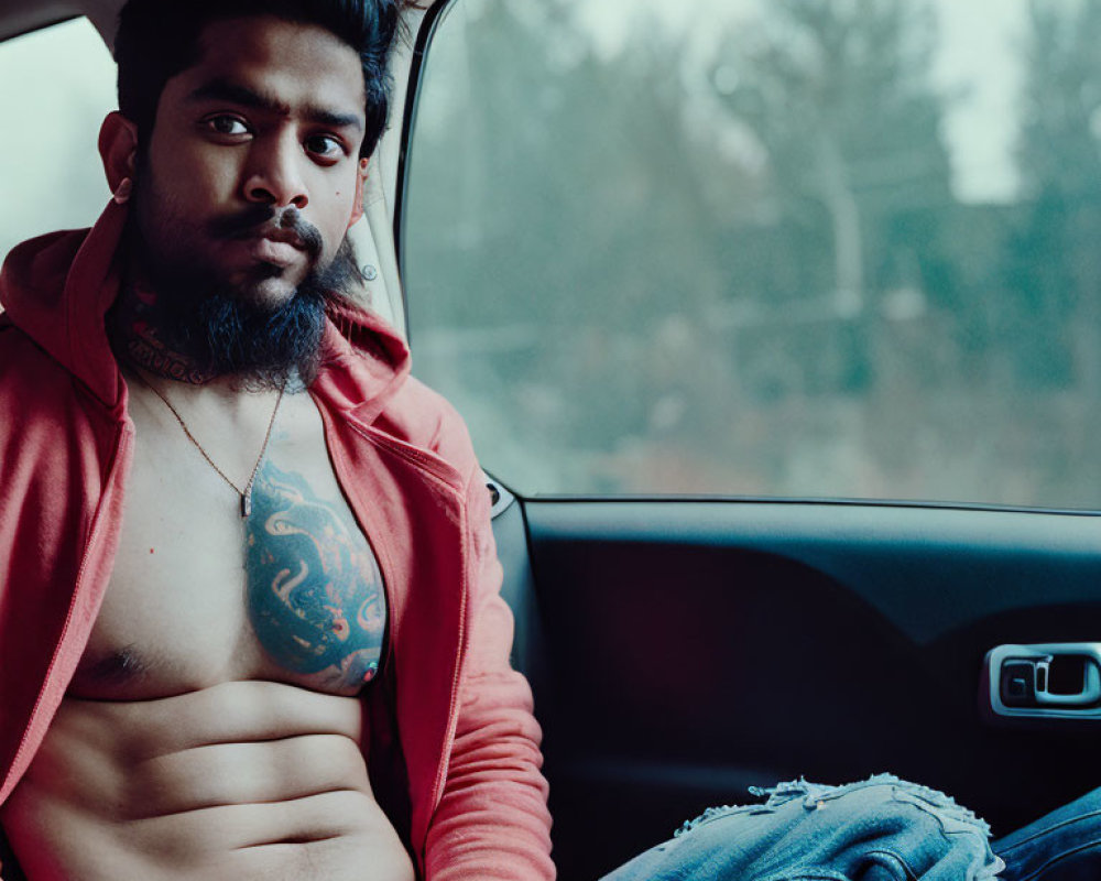 Tattooed man in red hoodie and ripped jeans sitting in car