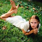 Person lying on stomach in grass with bare feet up, head on hands, intense expression