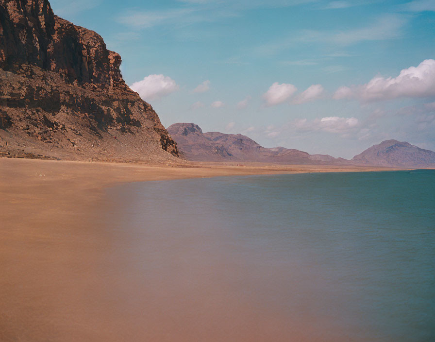 Tranquil beach scene with sandy shore, calm waters, mountains, and cloudy sky