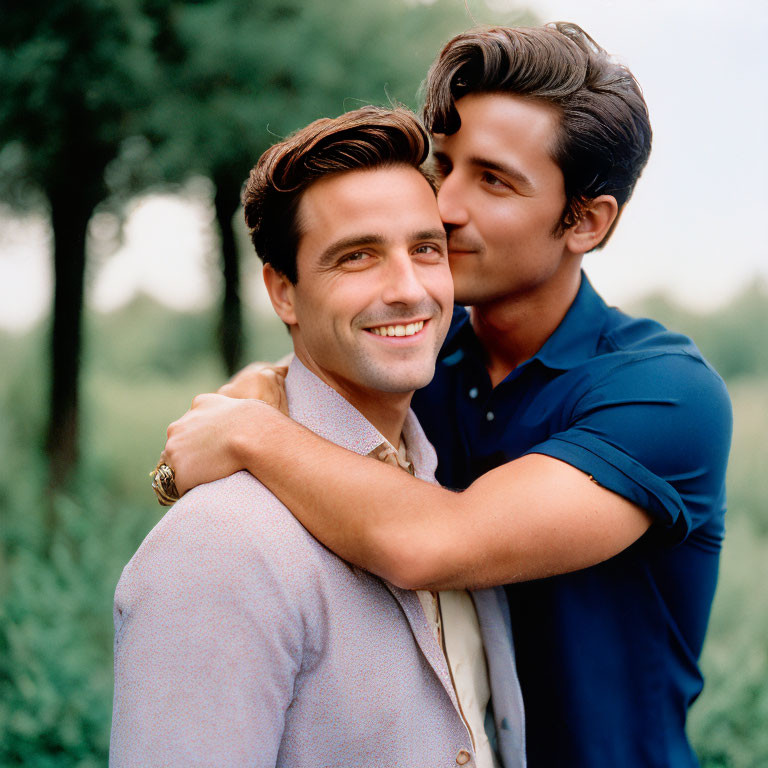 Two men embracing outdoors in smart casual attire, one kissing the other's cheek affectionately