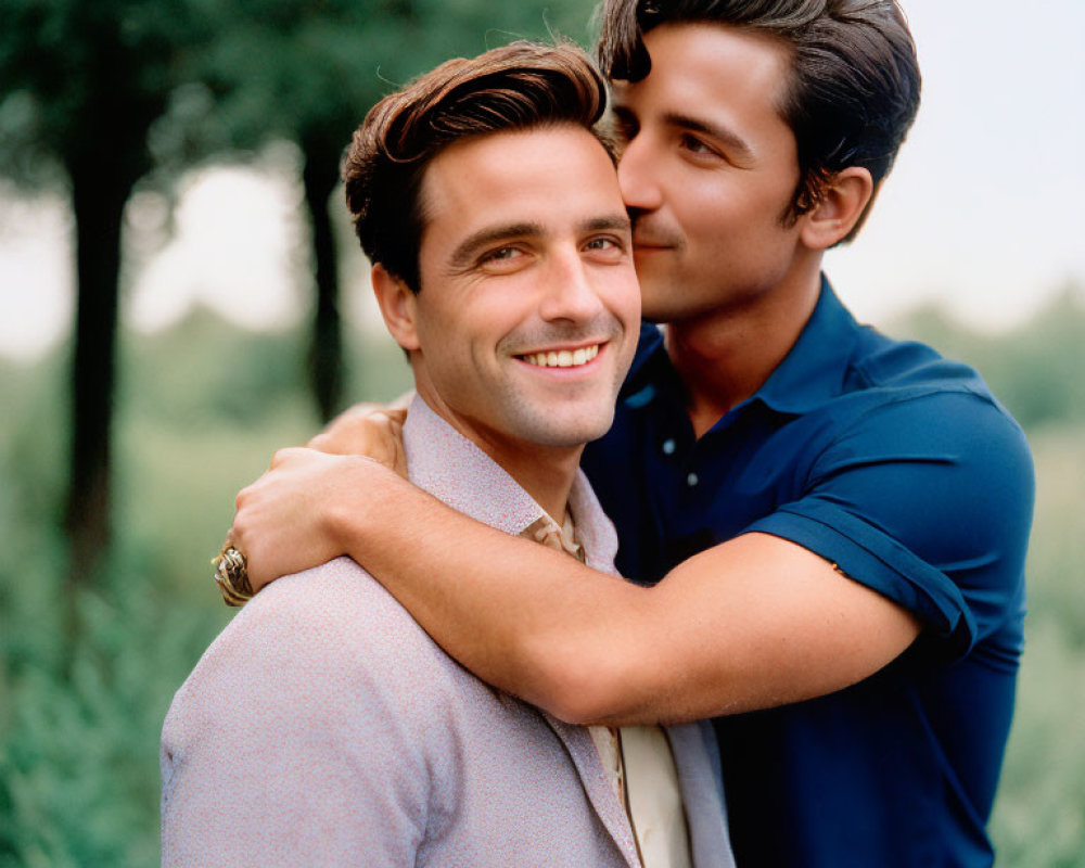 Two men embracing outdoors in smart casual attire, one kissing the other's cheek affectionately