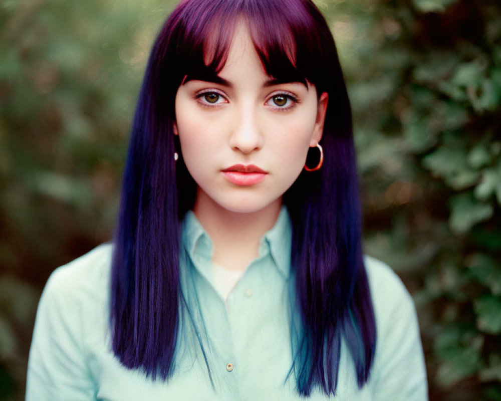 Purple-haired woman in mint-green blouse gazes at camera with blurred foliage background