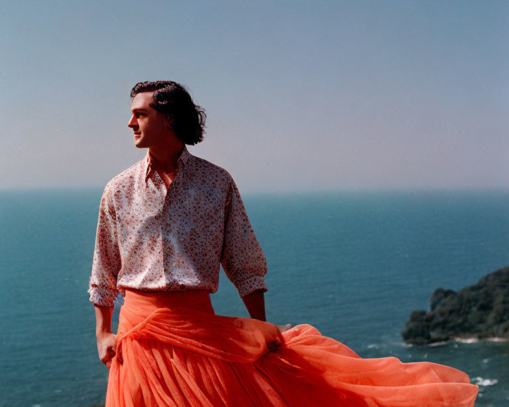 Person in patterned shirt and orange skirt gazes at sea from coastal overlook