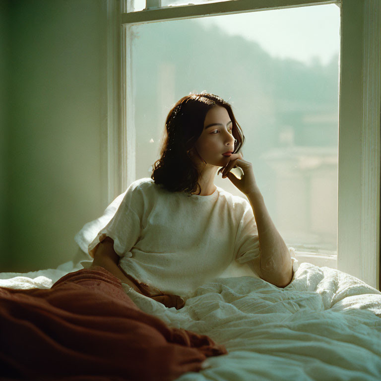 Pensive person sitting on bed by window in natural light