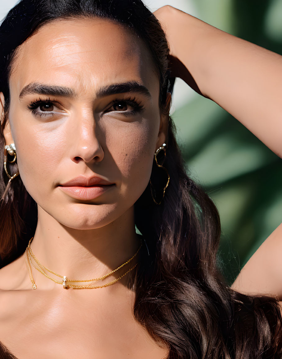 Portrait of woman with dark hair, hooped earrings, delicate necklace, green leaves backdrop.