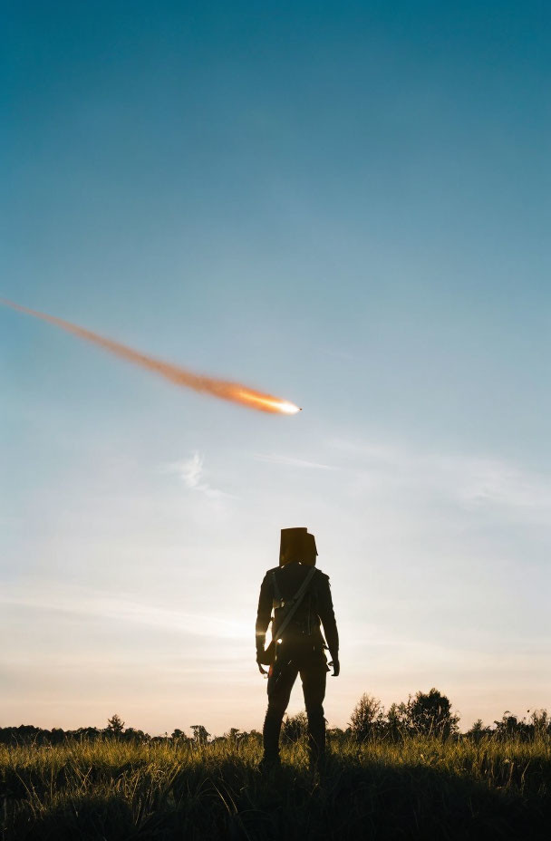 Figure in hat stands in field at sunset with meteor in sky